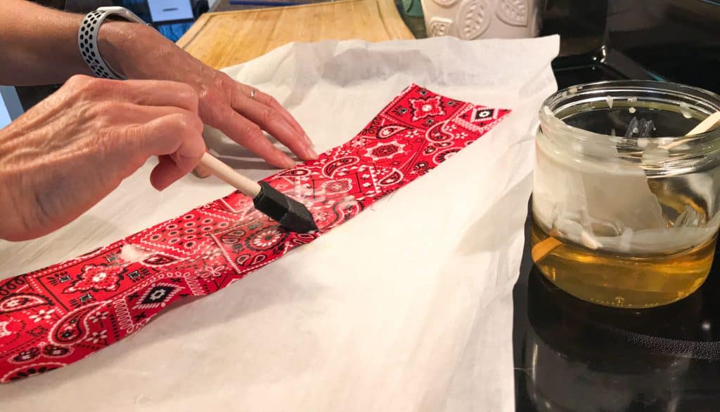 picture showing hand holding a foam craft brush and brushing on melted wax on a piece of red bandana fabric